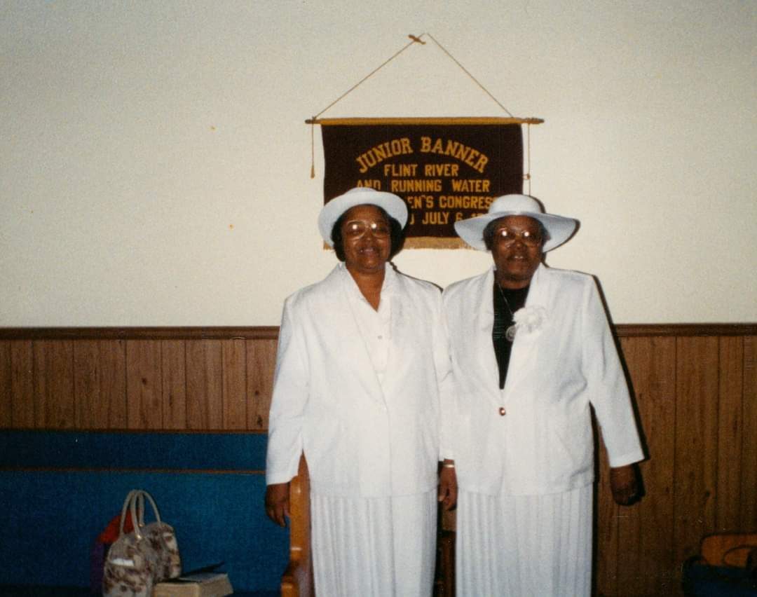 Two church mothers dressed in white with white hats, standing beneath a Flint River and Running Water Junior Banner