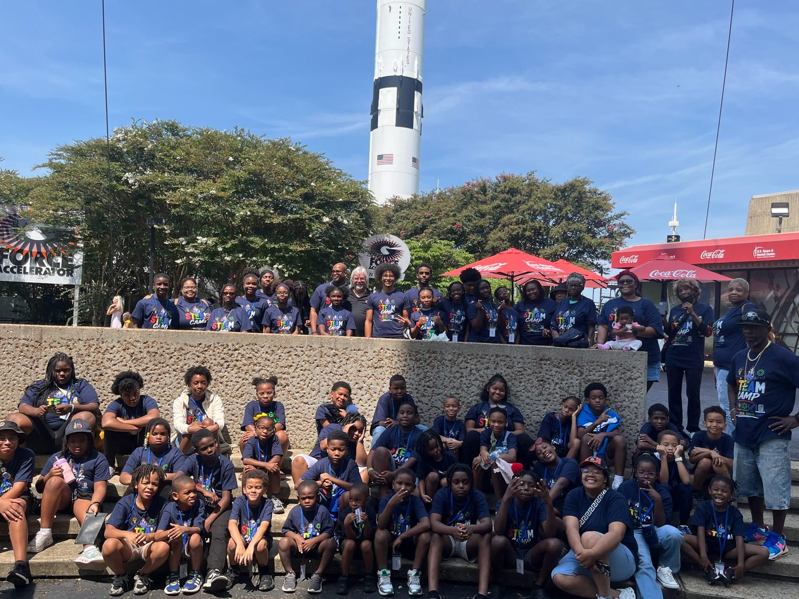 Dozens of STEAM Summer Camp kids and volunteers in matching navy camp shirts, posed in front of a Saturn rocket